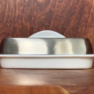 May include: A rectangular butter dish featuring a white ceramic base and a brushed stainless steel lid. The lid has a smooth, rounded top profile and sits securely within the recessed rim of the white ceramic tray, creating a clean, two-tone look.