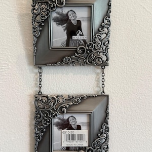 May include: A double hanging picture frame featuring two square metallic silver frames connected by small chains. Each frame has ornate scrollwork detailing on the top-left and bottom-right corners. The frames contain black and white photos.