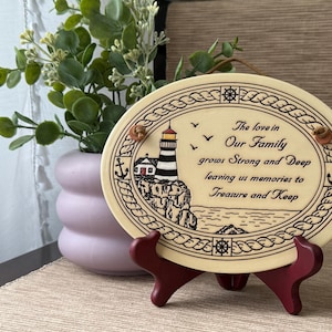 May include: An oval cream-colored plaque featuring a black line drawing of a lighthouse on a cliff, nautical rope borders, and the text: The love in Our Family grows Strong and Deep leaving us memories to Treasure and Keep, held by a dark red stand.