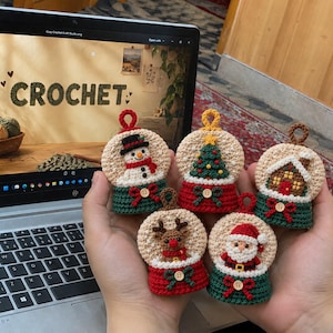 Può includere: Cinque piccoli ornamenti all'uncinetto a forma di palla di neve, ognuno con un personaggio o una scena festiva diversa: un pupazzo di neve, un albero di Natale, una casa di pan di zenzero, una renna e Babbo Natale, con un piccolo bottone.
