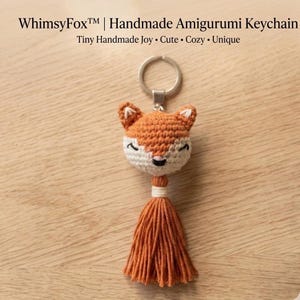 May include: A crochet fox head keychain with a metal ring, featuring orange and cream yarn, embroidered closed eyes, and a small black nose. Below the head is a long, orange yarn tassel tied with white thread. Text reads: WhimsyFox | Handmade Amigurumi Keychain Tiny Handmade Joy | Cute | Cozy | Unique