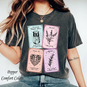 May include: A charcoal gray t-shirt featuring four rectangular graphic panels. The panels display black line art of a salt shaker, lavender, a rib cage, and a rosemary sprig, with text reading Always Throw Spilt Salt Over Your Left Shoulder, Plant For Luck, Fall In Love Whenever You Can, and Keep Rosemary By Your Garden Gate.