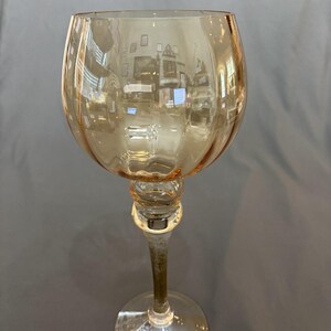 Puede incluir: Una copa de vino alta, de color ámbar, con un cuenco redondeado y un tallo delgado. La copa tiene una sección decorativa y bulbosa donde el tallo se une al cuenco. La base es circular y transparente. La copa refleja el entorno.