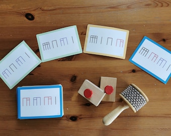 May include: A collection of educational musical rhythm cards with various patterns in black and red, alongside wooden rhythm blocks with red circles and a silver beaded maraca. The items are arranged on a wooden surface.