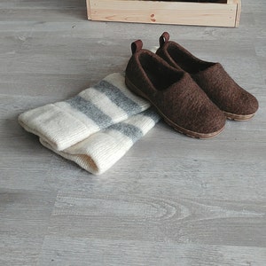 May include: Two brown wool slippers with a white and gray striped knitted scarf on a gray wood floor.  A white pitcher and potted plants are in wooden crates in the background.