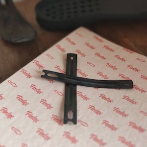 May include: Two black metal pieces shaped like a curved bracket, crossed over each other. The pieces are on a white surface with a repeating red pattern of the word "Fintex".