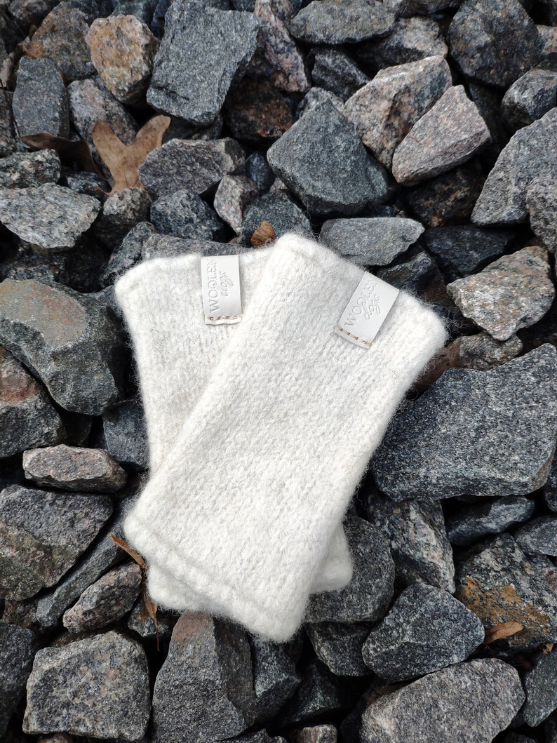May include: Two white knitted fingerless gloves with a white label that says "WOOLLEN 420F" on each glove. The gloves are on a bed of gray rocks.