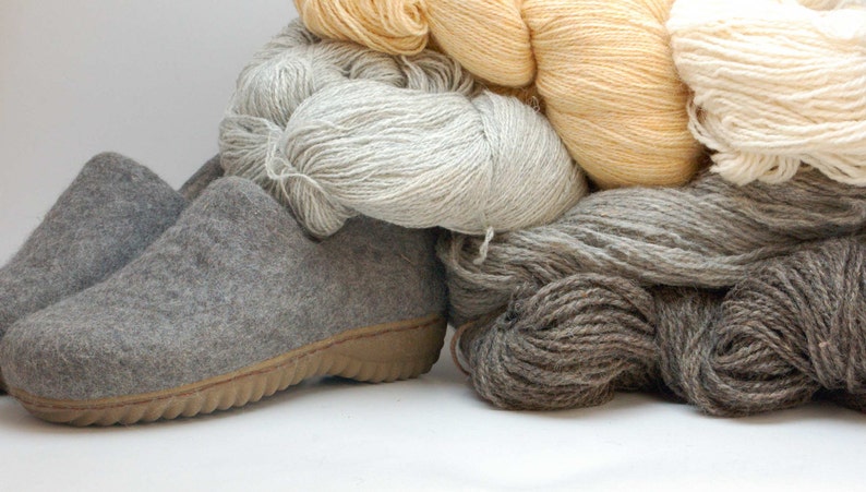 May include: Two gray felt slippers with a brown sole, sitting on a pile of yarn in various colors, including white, beige, and brown.