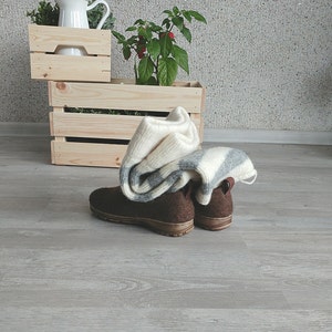 May include: Two pairs of brown wool slippers with a white and gray striped blanket on top of them. The slippers are on a light gray wood floor. There are two wooden crates in the background, one stacked on top of the other. The top crate has a white pitcher and green plants in it. The bottom crate has a green plant with red berries in it.