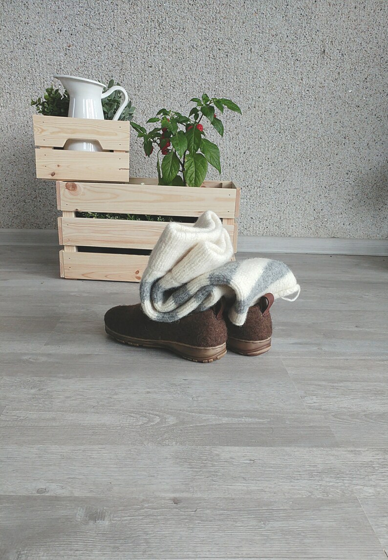 May include: Two pairs of brown wool slippers with a white and gray striped blanket on top of them. The slippers are on a light gray wood floor. There are two wooden crates in the background, one stacked on top of the other. The top crate has a white pitcher and green plants in it. The bottom crate has a green plant with red berries in it.