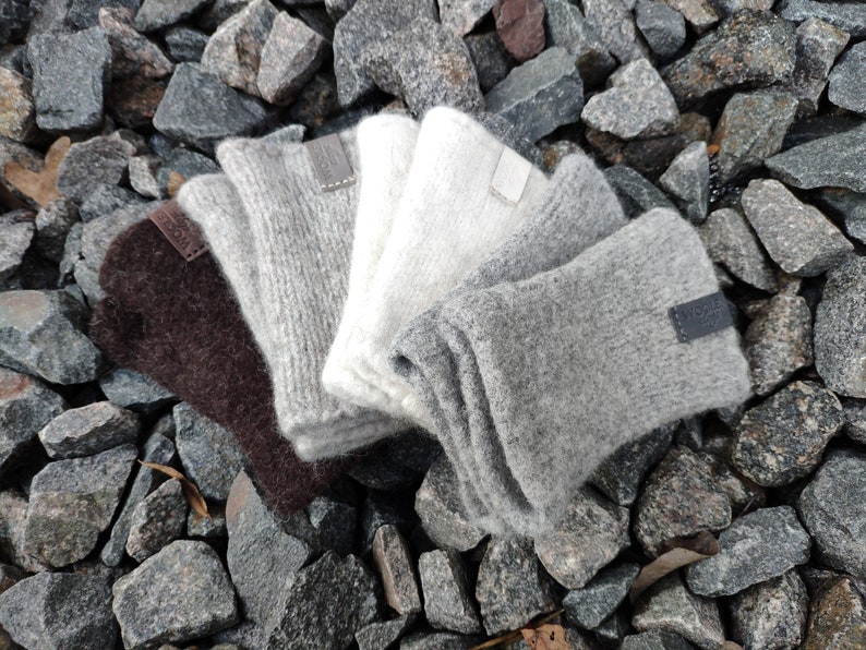 May include: Four pairs of fingerless gloves in different shades of gray, brown, and white. The gloves are made of a soft, fuzzy material and have leather labels with the word "Wool" on them.