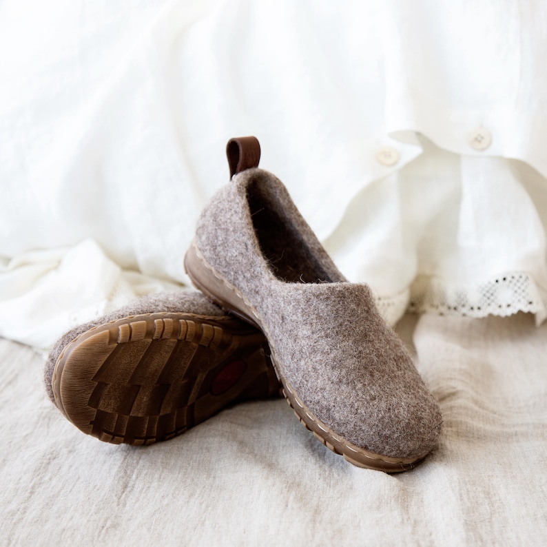 Peut inclure: Une paire de pantoufles en feutre marron avec une bordure en cuir marron. Les pantoufles sont sur une surface en tissu brun clair.