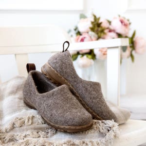 Peut inclure: Deux paires de pantoufles en feutre marron avec une bordure en cuir marron. Les pantoufles sont pos&eacute;es sur une surface en tissu marron clair.