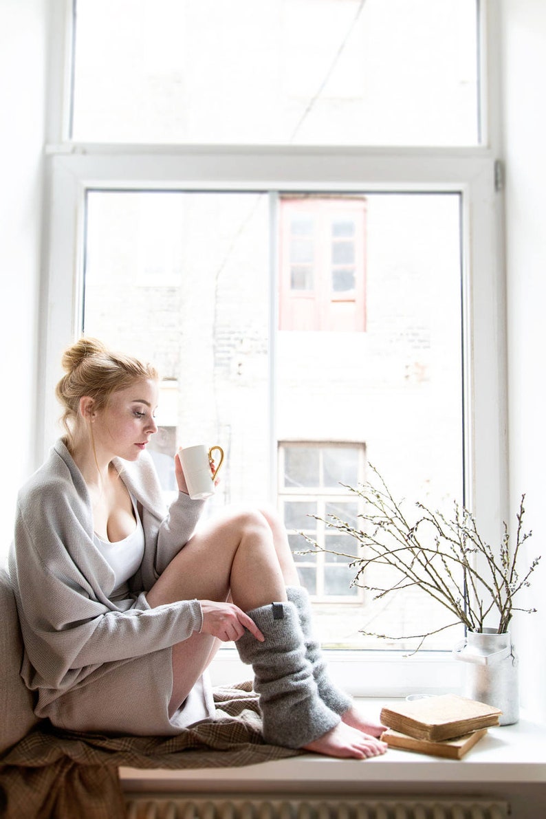 May include: A woman sits by a window, wearing a gray cardigan and gray leg warmers. She is holding a white mug with a gold handle. There are books and a vase with branches on the windowsill.