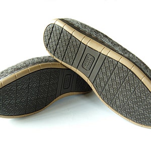 May include: A pair of gray wool slippers with a brown rubber sole. The soles have a textured pattern for grip. The slippers are designed for indoor use and provide comfort and warmth. The brand name is visible on the sole.