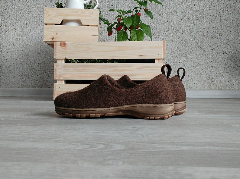 Peut inclure: Une paire de pantoufles en feutre marron avec une semelle en caoutchouc. Les pantoufles sont pos&eacute;es sur un sol en bois.