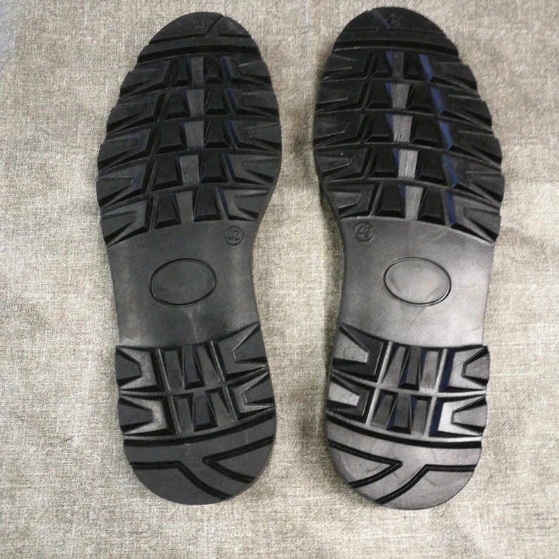 May include: Close-up of the black rubber soles of a pair of boots. The soles have a deep tread pattern for traction.