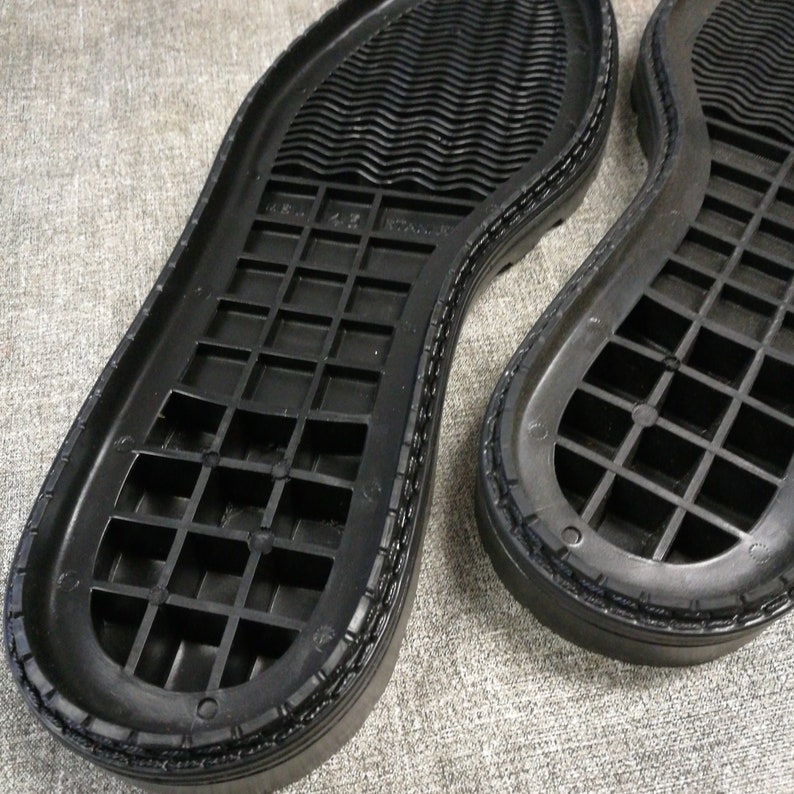 May include: Close-up of the black rubber soles of two shoes. The soles have a grid pattern and a textured edge.