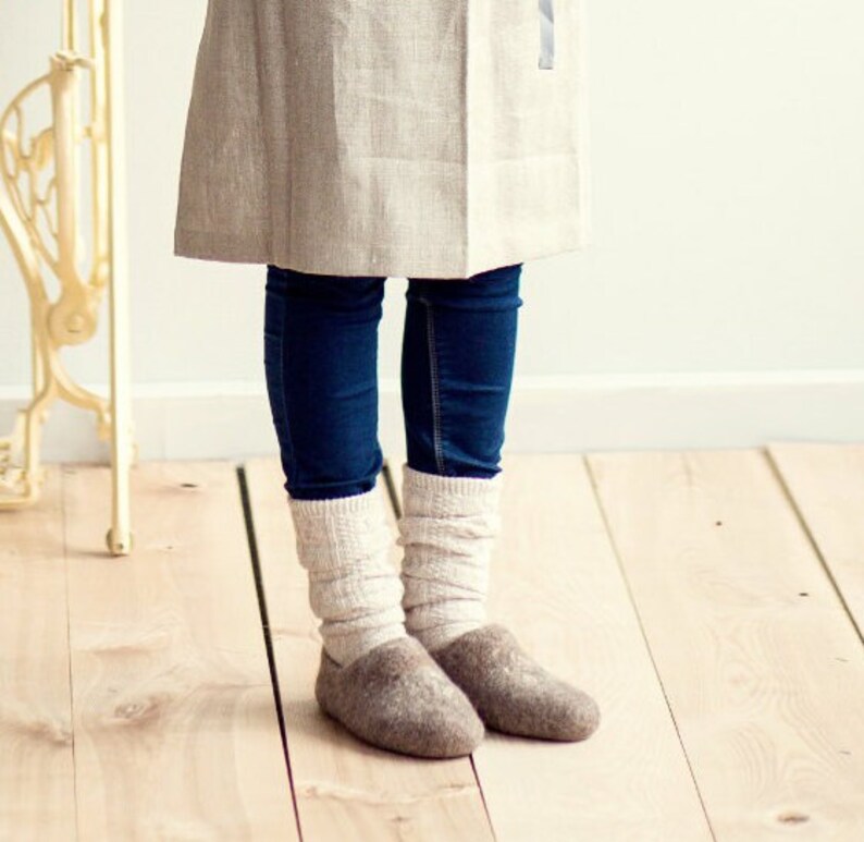 Pode incluir: Um par de pantufas de l&atilde; marrom com uma bainha de malha branca. As pantufas s&atilde;o usadas por uma pessoa que usa jeans azul e uma saia de linho marrom claro.