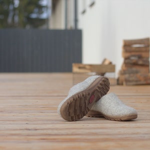 Peut inclure: Une paire de pantoufles en laine grise avec des semelles en caoutchouc brun. Les pantoufles sont sur une terrasse en bois.