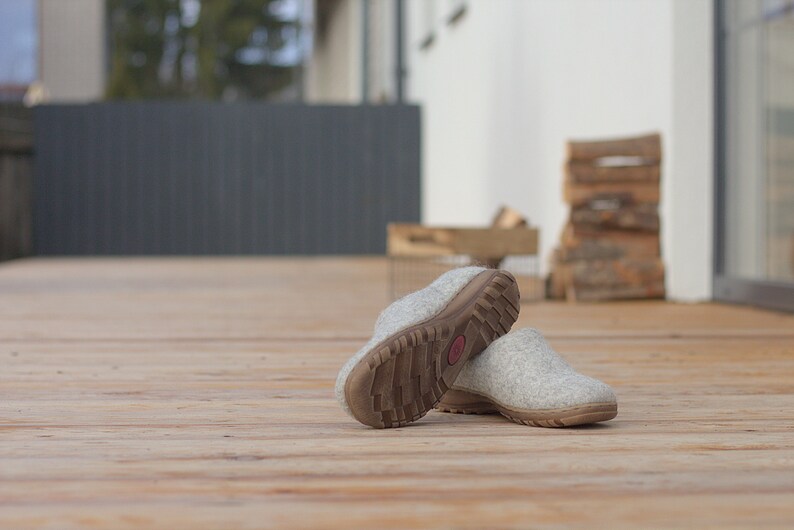 Peut inclure: Une paire de pantoufles en laine grise avec des semelles en caoutchouc brun. Les pantoufles sont sur une terrasse en bois.