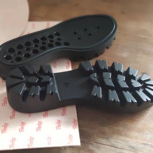 Puede incluir: Dos suelas de zapatos de goma negra. Una suela tiene un patr&oacute;n de banda de rodadura texturizado y la otra tiene un patr&oacute;n de peque&ntilde;os agujeros.