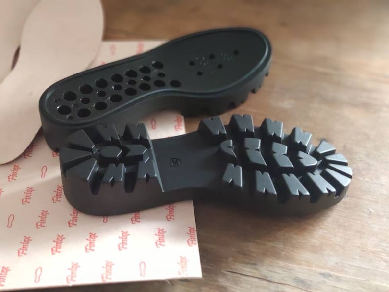 Puede incluir: Dos suelas de zapatos de goma negra. Una suela tiene un patr&oacute;n de banda de rodadura texturizado y la otra tiene un patr&oacute;n de peque&ntilde;os agujeros.