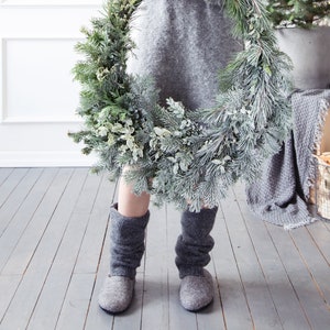 May include: A person wearing a gray sweater and gray felt boots is holding a large evergreen wreath decorated with frosted pine needles and eucalyptus.