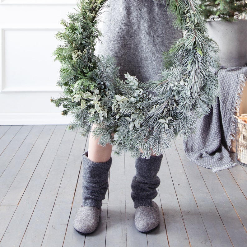 May include: A person wearing a gray sweater and gray felt boots is holding a large evergreen wreath decorated with frosted pine needles and eucalyptus.