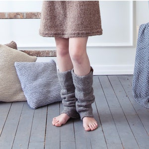 May include: A woman wearing a brown knitted dress and brown knitted leg warmers. Three knitted pillows in beige, brown, and gray are on the floor. A blue and white blanket is draped over a wooden box.