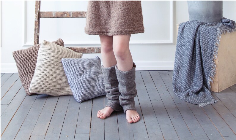 May include: A woman wearing a brown knitted dress and brown knitted leg warmers. Three knitted pillows in beige, brown, and gray are on the floor. A blue and white blanket is draped over a wooden box.