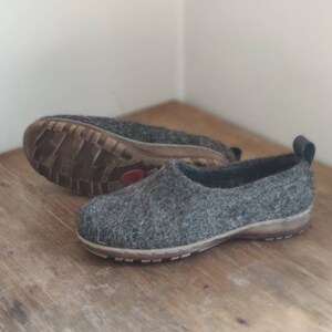 Peut inclure: Une paire de pantoufles en feutre gris avec des bordures noires et une semelle en caoutchouc marron. Les pantoufles sont sur une surface en bois.