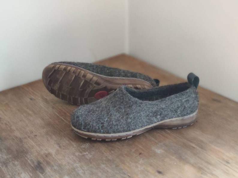 Peut inclure: Une paire de pantoufles en feutre gris avec des bordures noires et une semelle en caoutchouc marron. Les pantoufles sont sur une surface en bois.