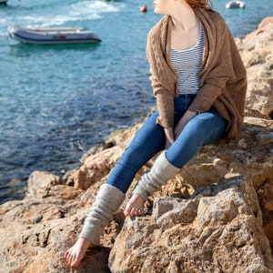 May include: A woman wearing a brown cardigan, blue jeans, and grey leg warmers sits on a rocky shore by the ocean.