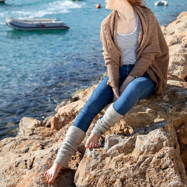 May include: A woman wearing a brown cardigan, blue jeans, and grey leg warmers sits on a rocky shore by the ocean.