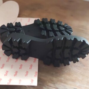 Puede incluir: Primer plano de las suelas de goma negra de un par de zapatos. Las suelas tienen un patr&oacute;n de banda de rodadura profundo.