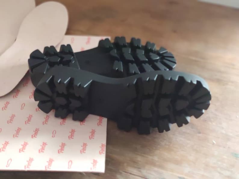 Puede incluir: Primer plano de las suelas de goma negra de un par de zapatos. Las suelas tienen un patr&oacute;n de banda de rodadura profundo.