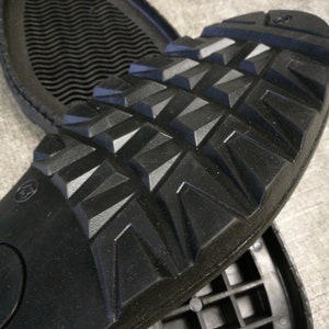 May include: Close-up of a black rubber sole with a textured diamond pattern. The sole is designed for traction and durability.