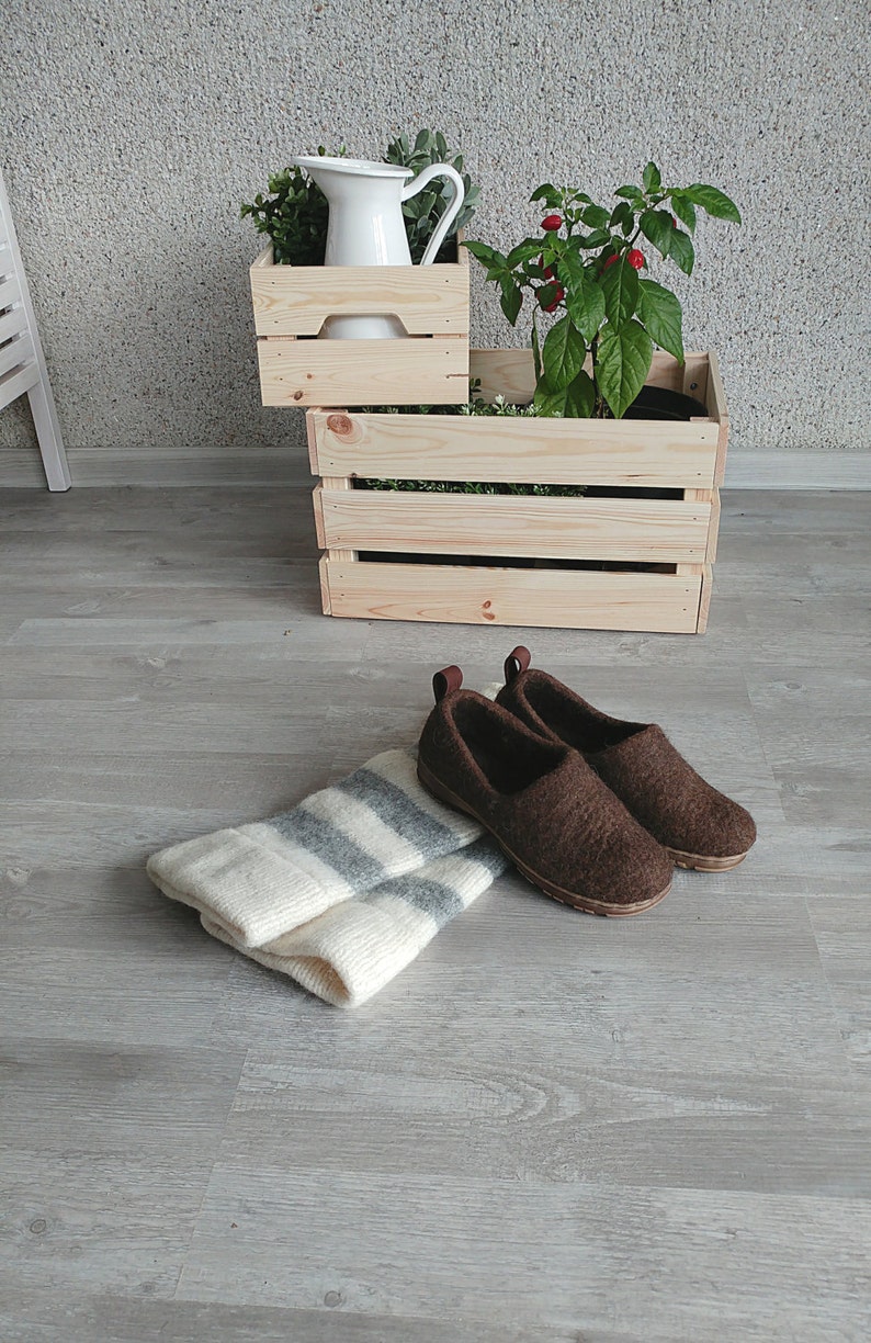 May include: Two brown wool slippers with a white and gray striped knitted scarf on a gray wood floor.  A white pitcher and potted plants are in wooden crates in the background.