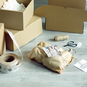 Pode incluir: Um pacote de papel castanho amarrado com barbante e um cart&atilde;o pequeno. O pacote est&aacute; rodeado de caixas de cart&atilde;o, um rolo de fita de papel castanho e um carretel de barbante. A fita tem uma etiqueta que diz "Wollen".