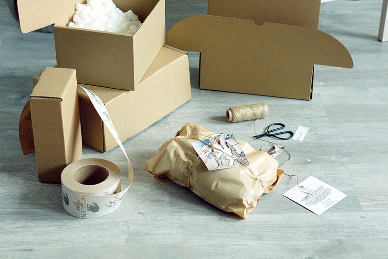 Pode incluir: Um pacote de papel castanho amarrado com barbante e um cart&atilde;o pequeno. O pacote est&aacute; rodeado de caixas de cart&atilde;o, um rolo de fita de papel castanho e um carretel de barbante. A fita tem uma etiqueta que diz "Wollen".