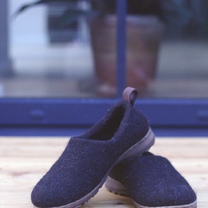 Peut inclure: Une paire de pantoufles en laine noire avec des semelles brunes. Les pantoufles sont sur une surface en bois.