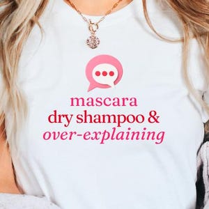 May include: White t-shirt with a pink speech bubble graphic containing three dots. Below the graphic, the words "mascara dry shampoo & over-explaining" are printed in pink. A gold necklace is visible at the top.