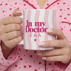 Peut inclure: Un mug en céramique avec un motif ondulé vertical rose clair et blanc. Le texte "In my Doctor ERA" est imprimé en lettres magenta grasses sur le devant, accompagné d'une petite icône de cœur magenta centrée sous le texte.