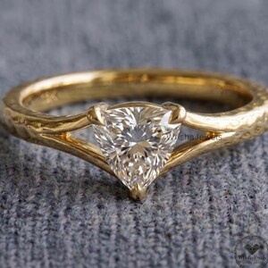 May include: A gold ring featuring a single trillion-cut clear gemstone set in a split shank band. The metal band has a textured, hammered finish and is stamped with 18K on the inner surface. The design centers on the triangular stone.