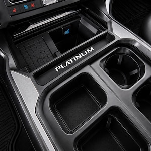 May include: A black car console with multiple compartments, including cup holders and storage areas. The word "PLATINUM" is displayed in white lettering. The console has a textured rubber mat and a charging port.
