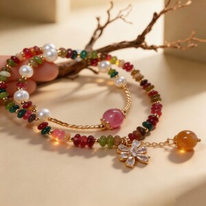 May include: A multi-strand bracelet featuring small, multicolored gemstone beads, white pearls, and gold-toned spacers. One strand includes a large pink bead, while the other features a dangling flower-shaped charm with clear stones and a yellow bead.