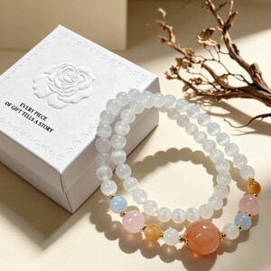 May include: Two beaded bracelets featuring translucent white spherical beads, accented with smaller pink, light blue, and orange beads, and one larger orange bead. The bracelets are positioned next to a white square gift box with a rose emblem.