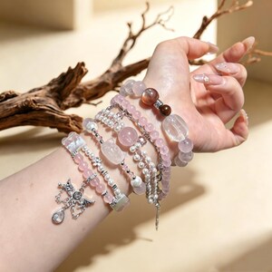 May include: A stack of multiple bracelets featuring light pink, clear, and brown beads, small white pearls, and silver-toned accents. One bracelet includes a silver-toned charm shaped like a winged heart with a dangling clear teardrop crystal.