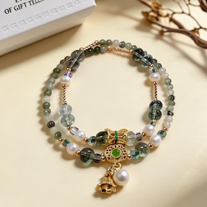 May include: A double-strand bracelet featuring green and translucent beads, white pearls, and gold-toned metal accents. The centerpiece includes a gold-toned filigree charm with a green inlay and a dangling white pearl, connected by gold-toned wire.